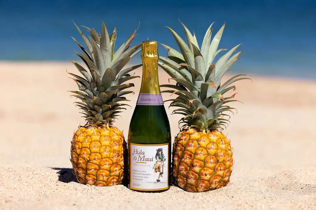 Bottle of Hula o Maui sparkling pineapple wine between two pineapples on the beach